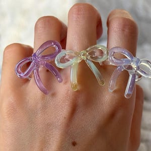 May include: Three transparent bow rings in shades of purple, yellow, and iridescent white are displayed on fingers. The rings have a delicate, whimsical design, perfect for adding a touch of playful elegance to any outfit.