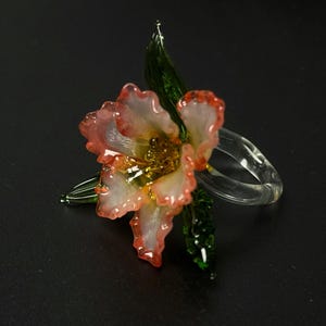 May include: A glass flower with pink and white petals, a yellow centre, and green leaves. The flower is attached to a clear glass ring. The flower is a decorative item.