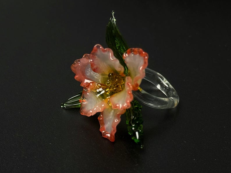 May include: A glass flower with pink and white petals, a yellow centre, and green leaves. The flower is attached to a clear glass ring. The flower is a decorative item.