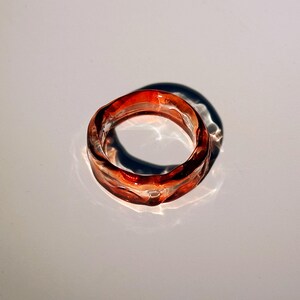 Pode incluir: Um anel transparente com um design ondulado. O anel &eacute; uma mistura de tons vermelhos e laranja, com um acabamento brilhante. O anel est&aacute; sobre uma superf&iacute;cie branca, projetando uma sombra.