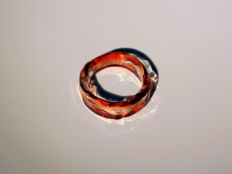 Pode incluir: Um anel transparente com um design ondulado. O anel &eacute; uma mistura de tons vermelhos e laranja, com um acabamento brilhante. O anel est&aacute; sobre uma superf&iacute;cie branca, projetando uma sombra.