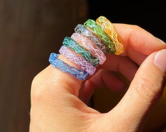 May include: A selection of colourful, transparent rings in shades of blue, purple, pink, green, and yellow. Each ring has a delicate, white, interwoven design. The rings are held in a hand, showcasing their vibrant colours and intricate patterns.