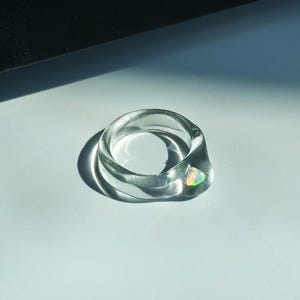 May include: A clear, chunky ring with a rounded design. The ring features a small, iridescent gemstone. The ring is made of a transparent material, and the light reflects off the surface, creating shadows.