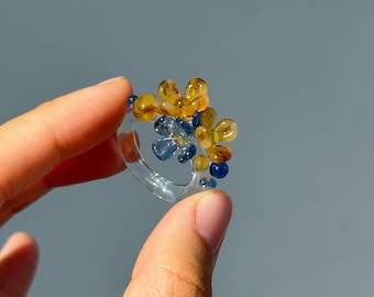 May include: A clear glass ring featuring yellow and blue floral decorations. The ring is held between fingers, displaying the intricate design against a grey backdrop. The ring's design is reminiscent of flowers.
