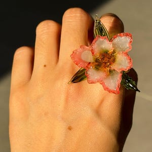 May include: A flower ring with a white and pink blossom design. The ring features a detailed flower with a gold centre and green leaves. The ring is a unique piece of jewellery.