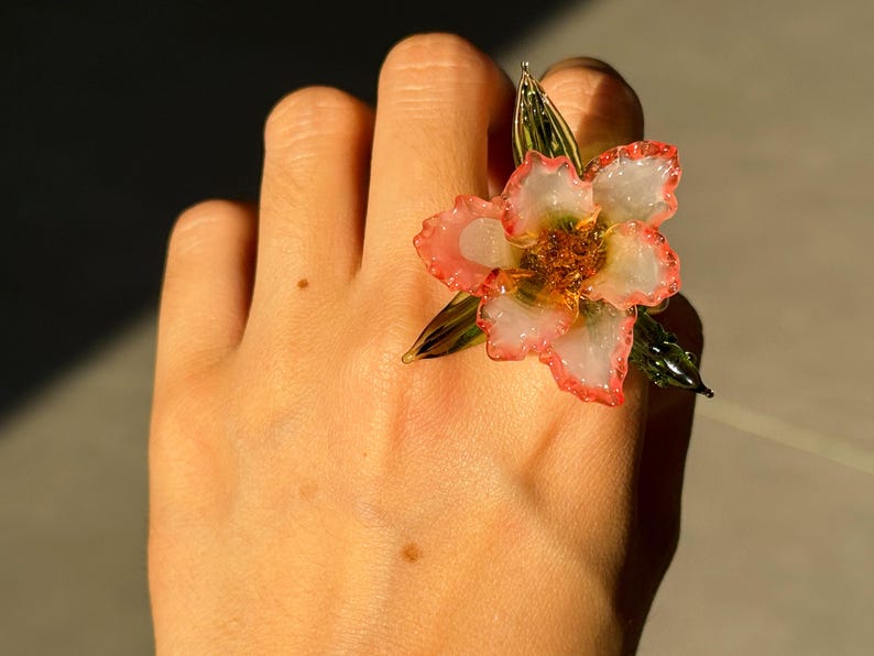 May include: A flower ring with a white and pink blossom design. The ring features a detailed flower with a gold centre and green leaves. The ring is a unique piece of jewellery.