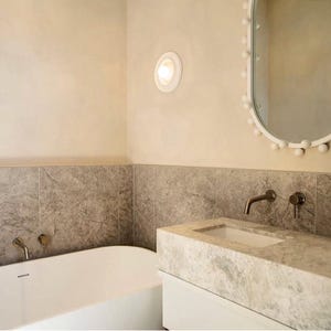 May include: A white bathroom with a bathtub, a sink, and a mirror with white pom-poms around the frame. The walls are a light beige colour and the floor is tiled in a light grey colour.