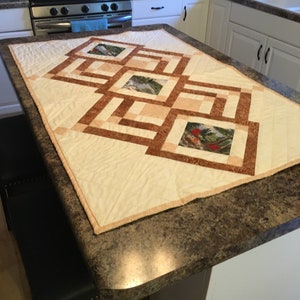 May include: A rectangular quilted table runner with a cream-colored background and a geometric design in shades of brown and tan. The design incorporates squares with nature scenes, including images of birds. The runner is placed on a kitchen countertop.