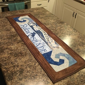 May include: A rectangular, wooden-framed table runner with a beach theme. The runner features blue and white geometric patterns and the text "Relax in the Shade" and "The Good Life". The runner is approximately 30 inches long.