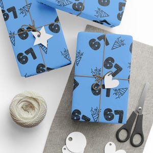 May include: Three wrapped gifts with blue paper featuring a black number 67 pattern and party-themed illustrations. Each gift is tied with twine and a white tag. A ball of twine, scissors, and blank tags are also present.