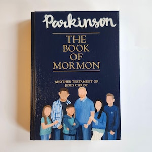 May include: A blue book with the title "The Book of Mormon" and the name "Parkinson" written in white cursive on the cover. The book also has the text "Another Testament of Jesus Christ" in gold lettering. A family portrait is painted on the cover, depicting a family of seven with blue clothing.