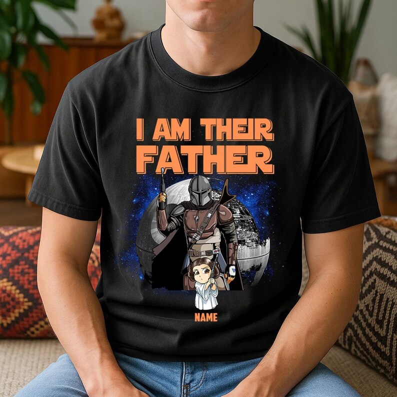 May include: Black t-shirt with the text "I AM THEIR FATHER" in orange. The shirt features a graphic of a character in armor holding a weapon, with a child figure below. A space scene is in the background.