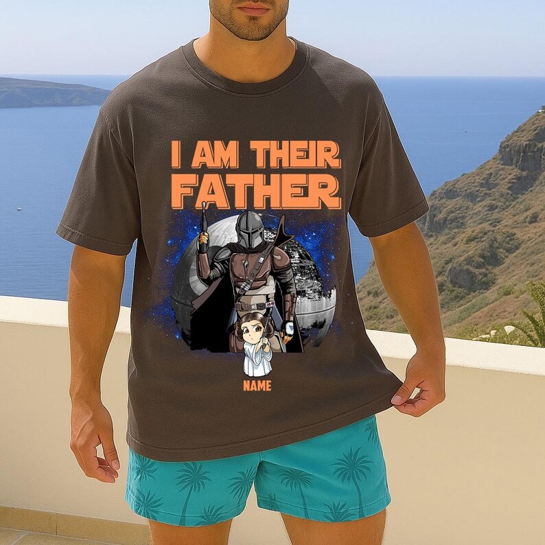 May include: Dark gray t-shirt with the text "I AM THEIR FATHER" in orange. The shirt features a graphic of a character in armor with a child, set against a starry background. The word "NAME" is below the child.