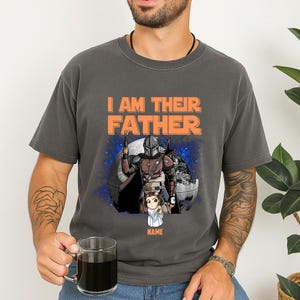May include: Dark gray t-shirt with the text "I AM THEIR FATHER" in orange. The shirt features a graphic of a Mandalorian character with a child, set against a space-themed background. The child's name is below the graphic.