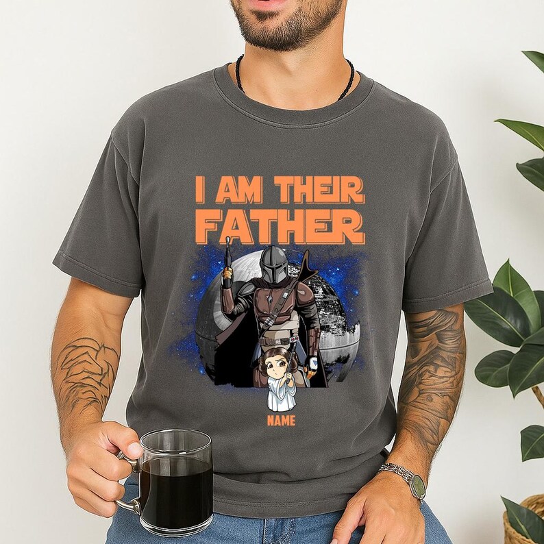 May include: Dark gray t-shirt with the text "I AM THEIR FATHER" in orange. The shirt features a graphic of a Mandalorian character with a child, set against a space-themed background. The child's name is below the graphic.