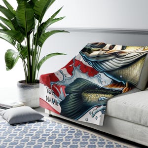 以下が含まれることがあります： ソファに掛けられた白いスローケット。緑、金、青の色合いで魚が詳細に描かれています。ブランケットには「YOUR NAME」の文字と赤と白の模様があります。
