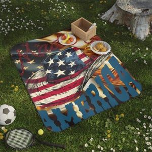 Può includere: Una coperta da picnic con un design a bandiera americana e la parola "AMERICA" in blu. L'immagine include un pallone da calcio, una racchetta da tennis e generi alimentari. La scena è ambientata sull'erba.