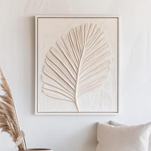 May include: A framed, off-white, textured wall art piece featuring a raised relief of a large palm leaf. The artwork is set against a white wall, with a neutral color palette. The frame is a simple, light-colored wood.