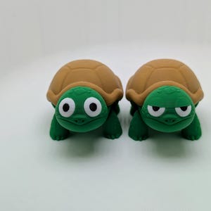 May include: Two green and brown turtle figurines with googly eyes. One turtle has a happy expression and the other has a grumpy expression.