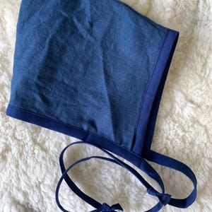 Könnte beinhalten: Ein blauer Hut aus Leinen mit dunkelblauer Einfassung und Bindebändern. Der Hut liegt auf einer weißen, flauschigen Oberfläche. Die Bänder werden unter dem Kinn gebunden.