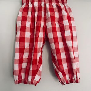 May include: Red and white checkered baby pants. The pants have an elastic waistband and gathered cuffs at the ankles. The fabric is a classic gingham pattern, perfect for a summer outfit.