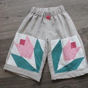 May include: Gray linen shorts with an elastic waistband and a pink button. The shorts feature a patchwork design of pink tulips with teal leaves and white polka dot backgrounds. A handmade garment.