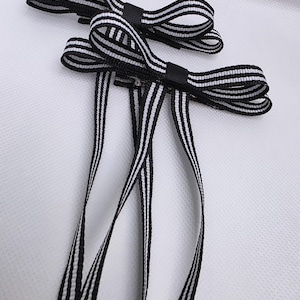 May include: Two black and white striped ribbon hair bows. Each bow has a small black center and long, striped ribbon tails. The bows are set against a white background.