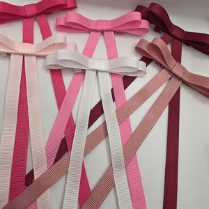May include: An assortment of ribbon hair bows in various shades of pink and white. The bows have a classic bow shape with long, vertical ribbon tails. The colors range from light pink to deep burgundy.