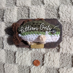 May include: A crocheted fish mounted on a wooden plaque, with the text "Pattern Only". The fish features green, white, and tan yarn, with a button eye. A penny is next to the plaque for scale. The background is a textured, checkered pattern.
