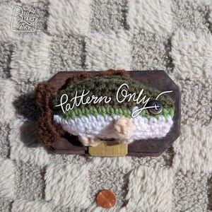 May include: Crocheted fish mounted on a wooden plaque. The fish features green, white, and brown yarn, with a button eye. The plaque has a brass-colored bracket. The text "Pattern Only" is written on the fish.