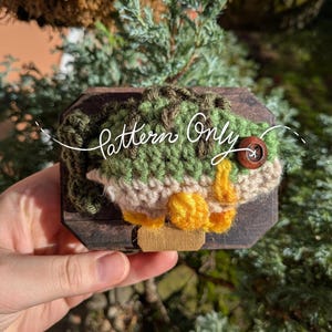 May include: A handmade crochet fish in shades of green, tan, and yellow, mounted on a dark wooden plaque. The fish has a button eye and is labeled "Pattern Only". The piece is held against a backdrop of green foliage.
