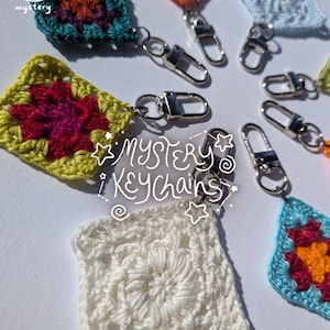 May include: A collection of colorful, crocheted square keychains. Each keychain features a unique floral design in various colors, including teal, orange, and yellow, with silver-tone metal clasps. The text "MYSTERY KEYCHAINS" is written on the image.