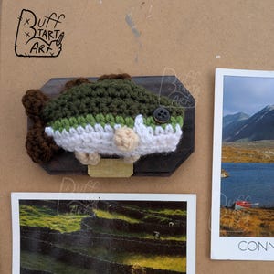Op de afbeelding: Een handgemaakte gehaakte vis gemonteerd op een donkere houten plaat. De vis heeft groen, wit en bruin garen, met een knoopoog en bruine vinnen. Het kunstwerk wordt getoond met twee landschapsfoto's.