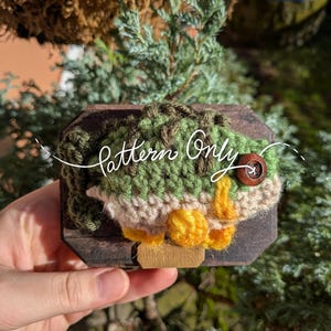 Puede incluir: Un diseño de pez tejido a ganchillo en tonos verdes, beige y amarillo, montado sobre una pequeña placa de madera. El texto "Pattern Only" está escrito en la imagen. Un botón de madera sirve como ojo.