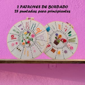 Puede incluir: Dos bastidores de bordado con tela blanca y puntadas de bordado de colores. Los bastidores están etiquetados con "2 PATRONES DE BORDADO 28 puntadas para principiantes".
