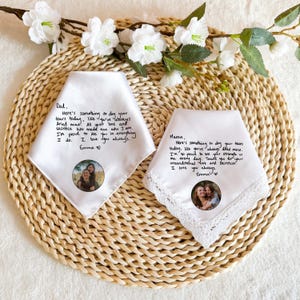 May include: Two white handkerchiefs with handwritten messages and photos. One says "Dad" and the other says "Mama". Both have a photo of a person with a child. The handkerchiefs are on a woven mat with white flowers.