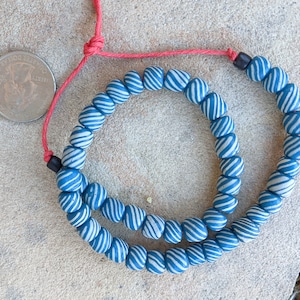 May include: A strand of blue and white striped beads, strung on a red cord. The beads are oval-shaped and have a spiral design. The cord is tied at the top and bottom with a black bead. The beads are displayed on a light brown surface.