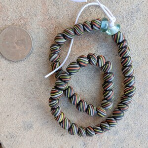 May include: Two black, orange, and white striped beaded bracelets. The bracelets are connected by a white string. The bracelets are on a light brown surface.