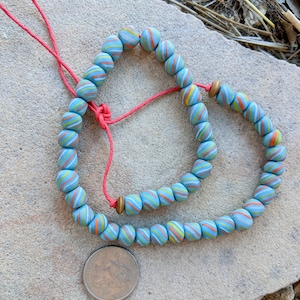 May include: A blue and white beaded necklace with orange and yellow stripes. The necklace is strung on a red cord and has a small gold bead accent.