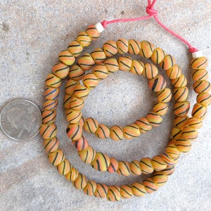 May include: A strand of yellow and orange glass beads with black and red stripes. The beads are strung on a red cord and are approximately the size of a US quarter.