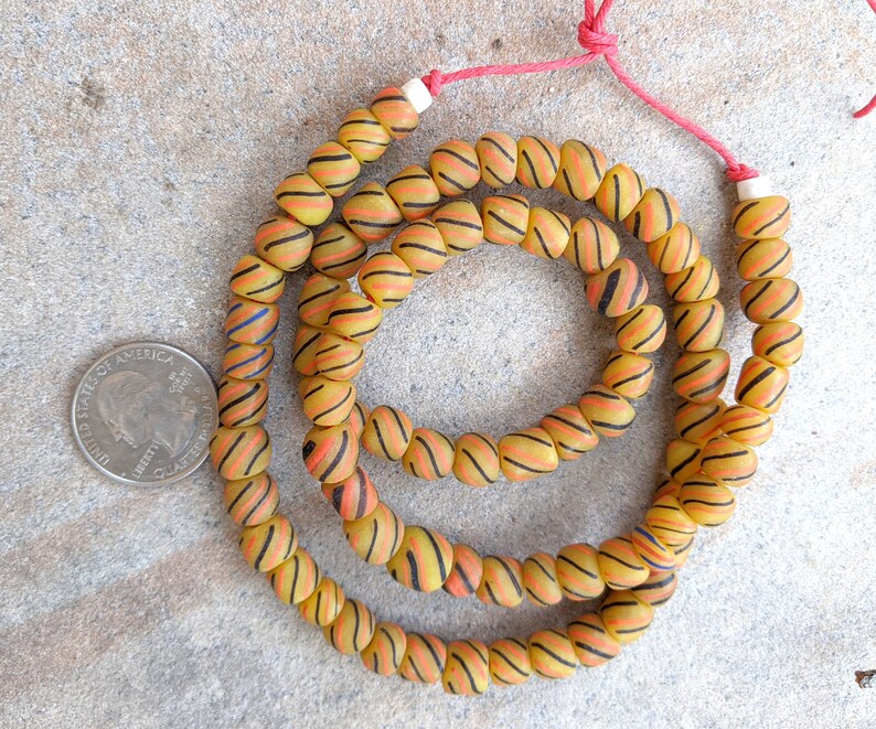 May include: A strand of yellow and orange glass beads with black and red stripes. The beads are strung on a red cord and are approximately the size of a US quarter.
