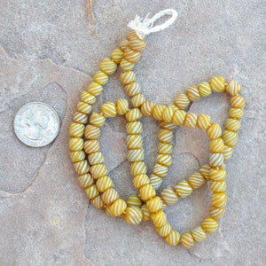May include: A strand of yellow and white striped glass beads. The beads are round and have a slightly irregular shape. The beads are strung on a white cord.