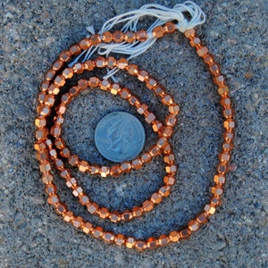 May include: A strand of copper-colored hexagonal beads, approximately the size of a US quarter, on a gray textured surface.