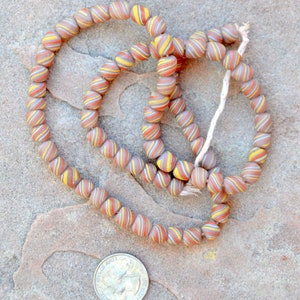 May include: A strand of brown, yellow, and white glass beads with a swirl pattern. The beads are strung on a white cord and are arranged in a circular shape.