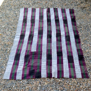 May include: A woven rug with a striped pattern in shades of white, purple, green, and black. The rug is made of a soft, textured fabric and has a fringed edge.