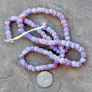 Puede incluir: Cadenas de cuentas de vidrio con base blanca y rayas en espiral rojas y azules. Las cuentas están ensartadas con un hilo blanco. Una moneda está al lado de las cuentas para la comparación de tamaño.