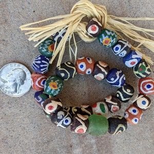 Puede incluir: Un collar de cuentas de colores con diferentes patrones y colores, incluyendo rojo, azul, verde y negro. Las cuentas están enhebradas en un cordón de fibra natural con un borla.