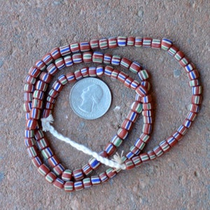 May include: A strand of red, white, blue, and green glass beads. The beads are small and cylindrical, and they are strung on a white cord. The beads are arranged in a pattern of alternating colors. The beads are about the size of a US quarter.