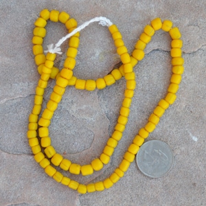 Puede incluir: Una hebra de cuentas de vidrio amarillo brillante, enhebradas en un cordón blanco. Las cuentas son pequeñas y cuadradas, y la hebra es lo suficientemente larga como para usarse como collar.