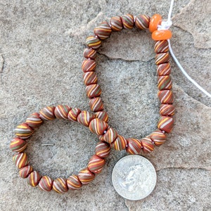 May include: A brown, orange, and white striped glass bead necklace with a silver coin charm. The necklace is strung on a white cord.